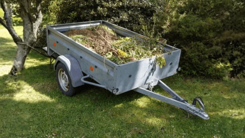 A grey colored dump trailer is parked in a garden and is loaded with dead shrubs