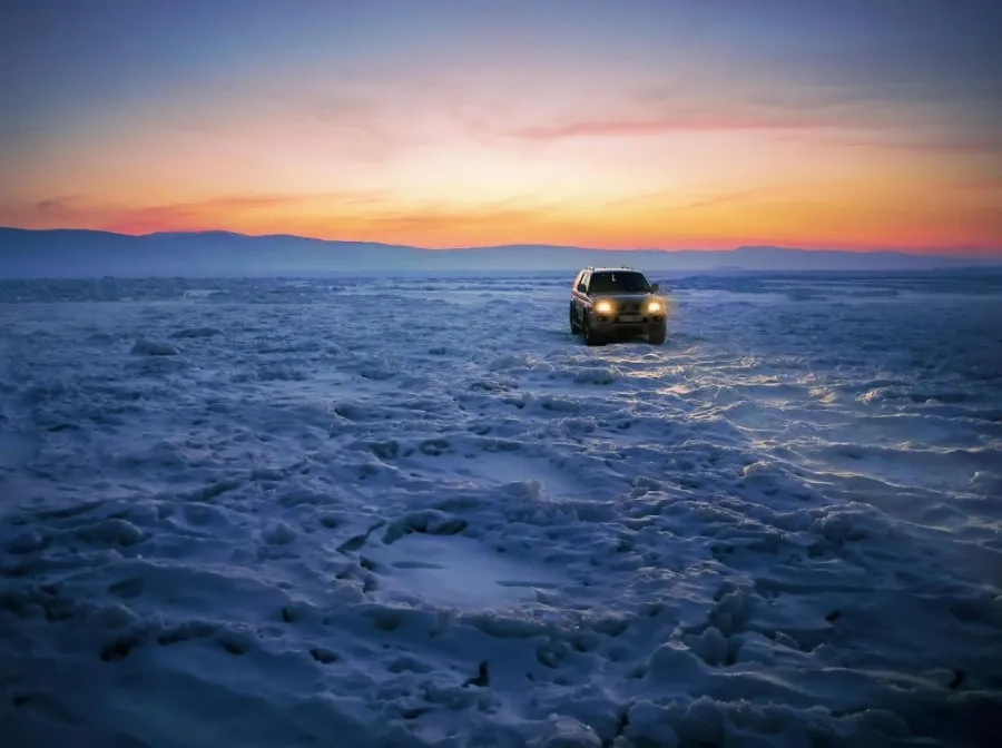 Ein Auto fährt während eines orangefarbenen Sonnenuntergangs über eine breite, ebene Schneedecke.