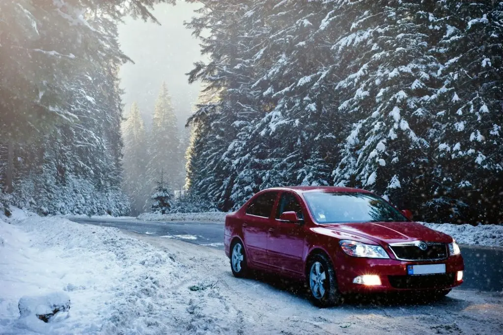 Sicherheitstipps für das Autofahren im Winter. Ein Auto fährt auf einer Straße durch einen dichten, schneebedeckten Kiefernwald. 