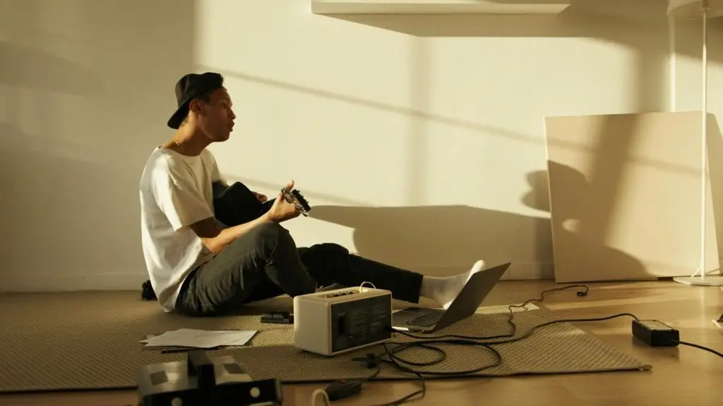 A man playing a guitar next to a laptop and a compact portable power station on the floor.