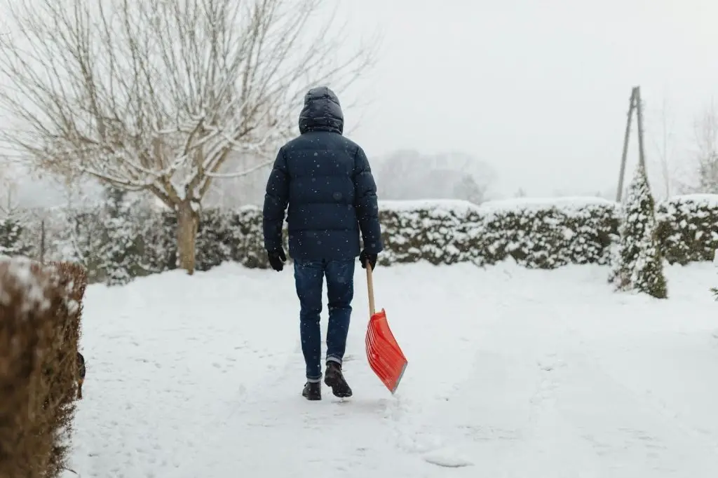 A man in winter gear holding a snow shovel, standing on a snow-covered driveway 