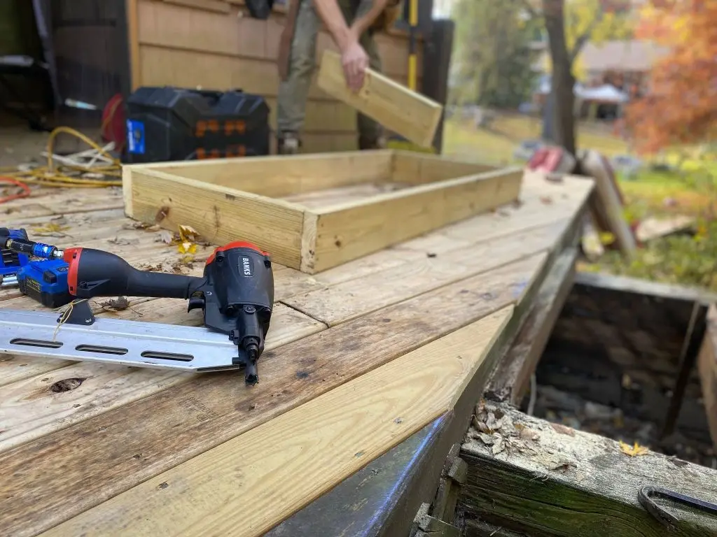 A man is assembling a wooden cold frame at the backyard of a house.