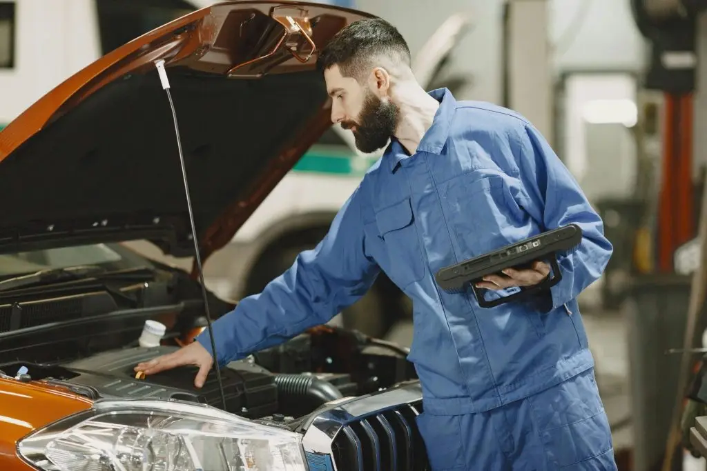 A man in a garage making a DIY car maintenace checklist while standing next to an open car hood.
